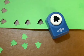 A blue shaped punch cutting small tree shapes from green paper.